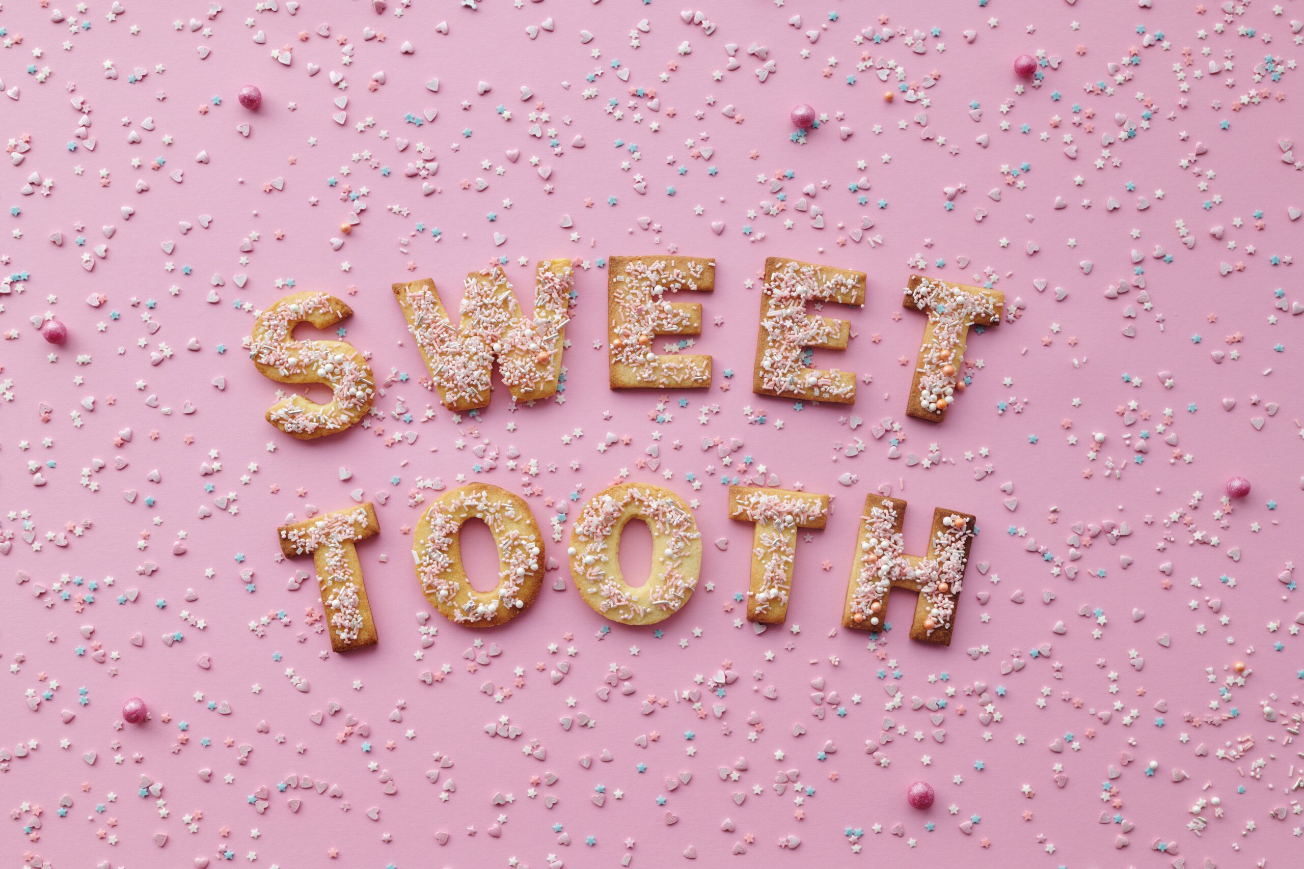 Flat lay of 'Sweet Tooth' cookies with sprinkles on a pink background. Perfect for dessert lovers.